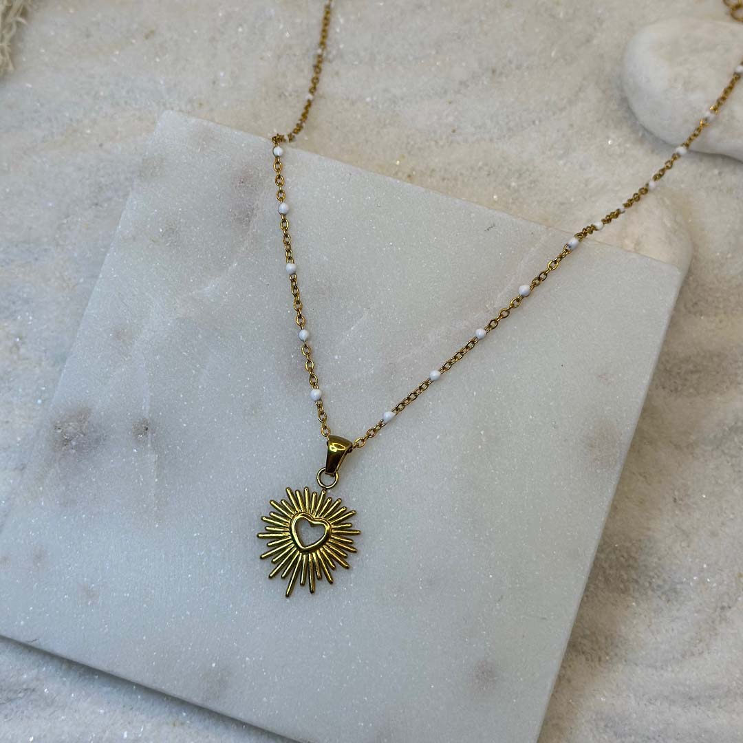 Un magnifique collier en acier inoxydable "Solia" de couleur Or, le bijou possède un pendentif de forme ronde avec un coeur au centre, ce bijou est posé sur une plaque de marbre blanche qui elle même est posé sur du sable blanc qui fait office de fond d'image.