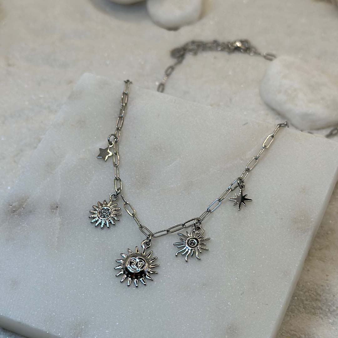 Un magnifique collier en acier inoxydable "Soline" de couleur argent, ce bijou possède plusieurs charms : 3 soleils de forme différentes ainsi qu'une étoile, le bijou est posé sur une plaque de marbre blanche qui elle même est posé sur du sable blanc qui fait office de fond d'image.