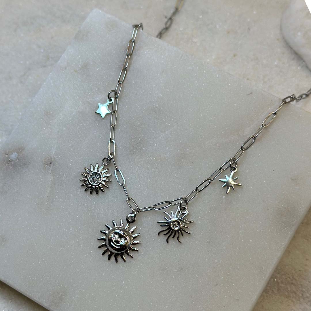 Un magnifique collier en acier inoxydable "Soline" de couleur argent, ce bijou possède plusieurs charms : 3 soleils de forme différentes ainsi qu'une étoile, le bijou est posé sur une plaque de marbre blanche qui elle même est posé sur du sable blanc qui fait office de fond d'image.