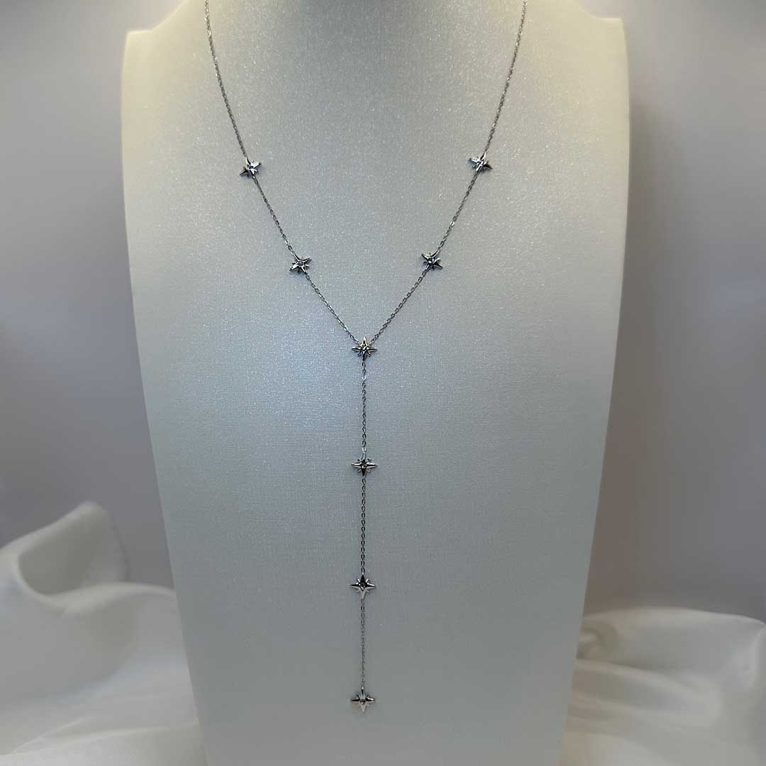 Un magnifique collier en acier inoxydable "Stary" de couleur argent, ce collier est en forme Y avec plusieurs étoiles de couleur argent tout le long de ce dernier, le bijoux est posé sur un portant mannequin buste blanc avec un draps en satin blanc a la base de ce portant