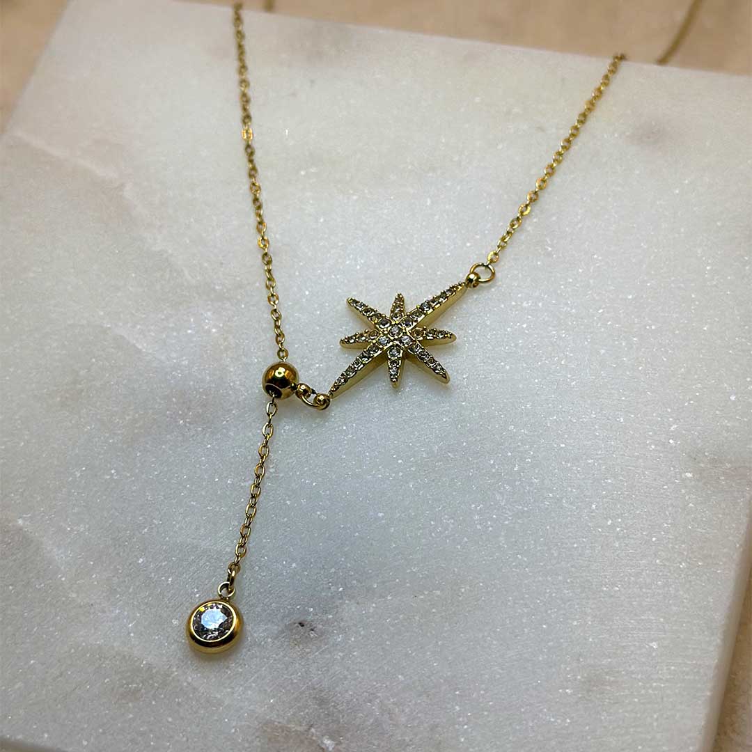 Un magnifique collier en acier inoxydable "Stella" de couleur or, ce bijou possède un charm en forme d'étoile incrusté de diamant ainsi qu'un strass en pendentif, ci bijou est posé sur une plaque de marbre blanche qui elle même est posé sur du sable blanc qui fait office de fond d'image.