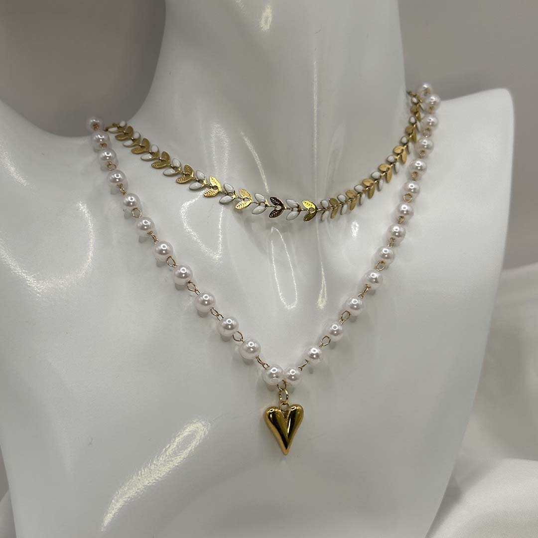 Deux magnifiques colliers en acier inoxydable sont posés sur un buste blanc brillant. Un collier "taina" est composé d'enchaînement de feuilles de couleur doré et blanc. Le 2e collier "Jane" est composé de perles blanches nacrées avec un pendentif en coeur de couleur or. C'est une composition imaginée par nos soins. Le buste est posé sur un fond blanc et un tissu en satin blanc.