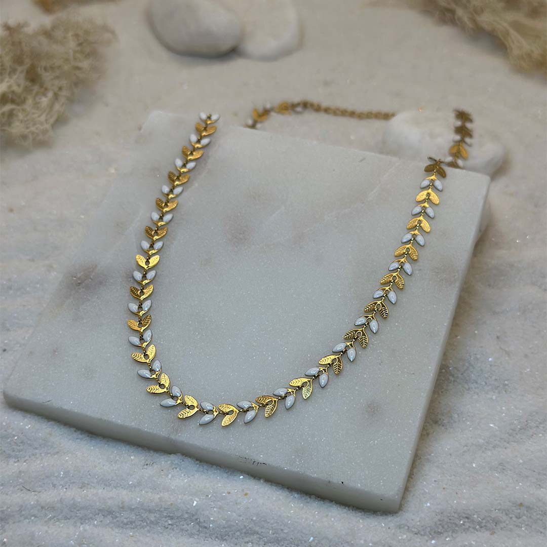 Un magnifique collier en acier inoxydable "Taina" de couleur Or, le bijou possède une chaine en forme de fleur alternant les couleurs or et blanc, ce bijou est posé sur une plaque de marbre blanche qui elle même est posé sur du sable blanc qui fait office de fond d'image.