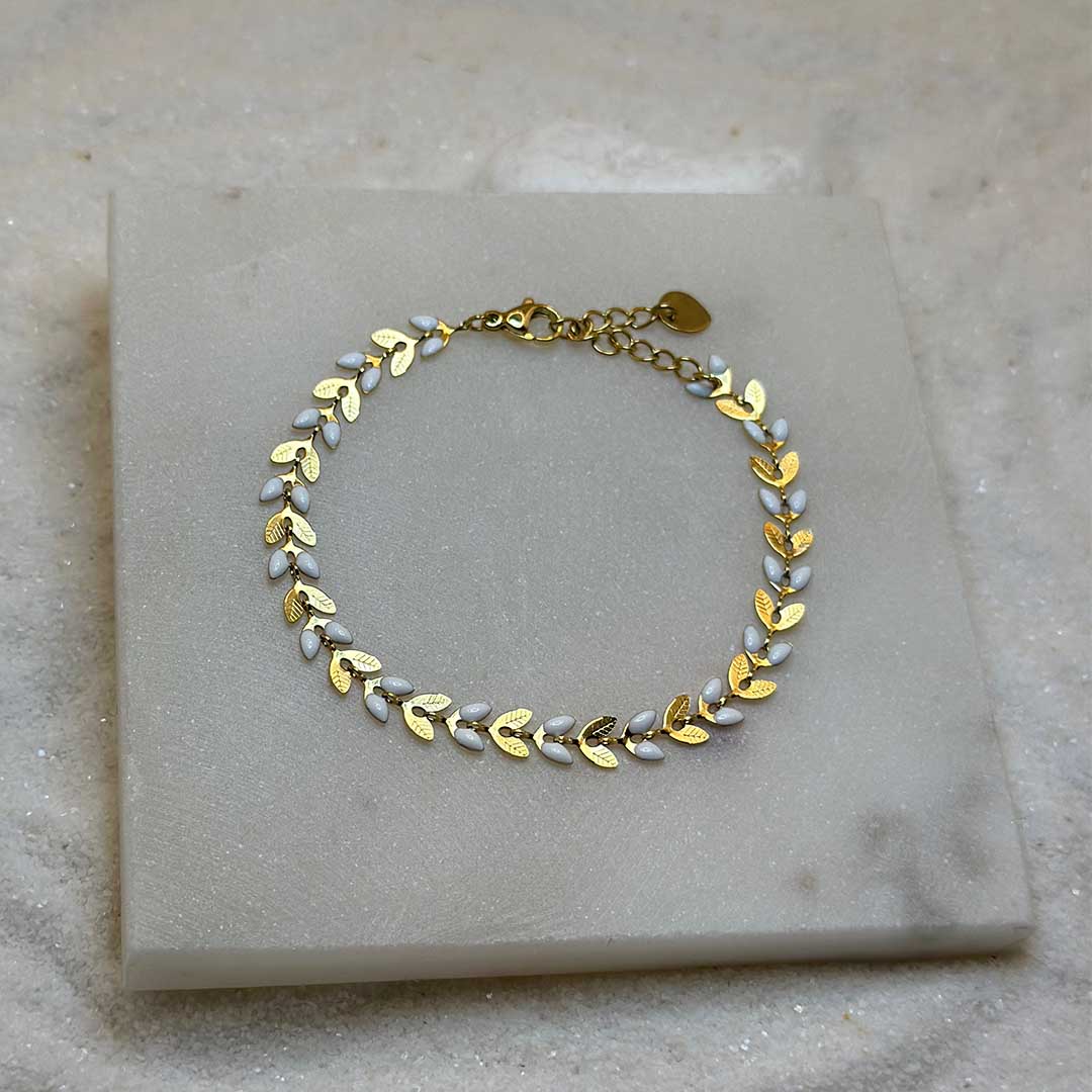 Un magnifique bracelet en acier inoxydable "Taina" de couleur Or, le bijou possède une chaine en forme de fleur alternant les couleurs or et blanc, ce bijou est posé sur une plaque de marbre blanche qui elle même est posé sur du sable blanc qui fait office de fond d'image.