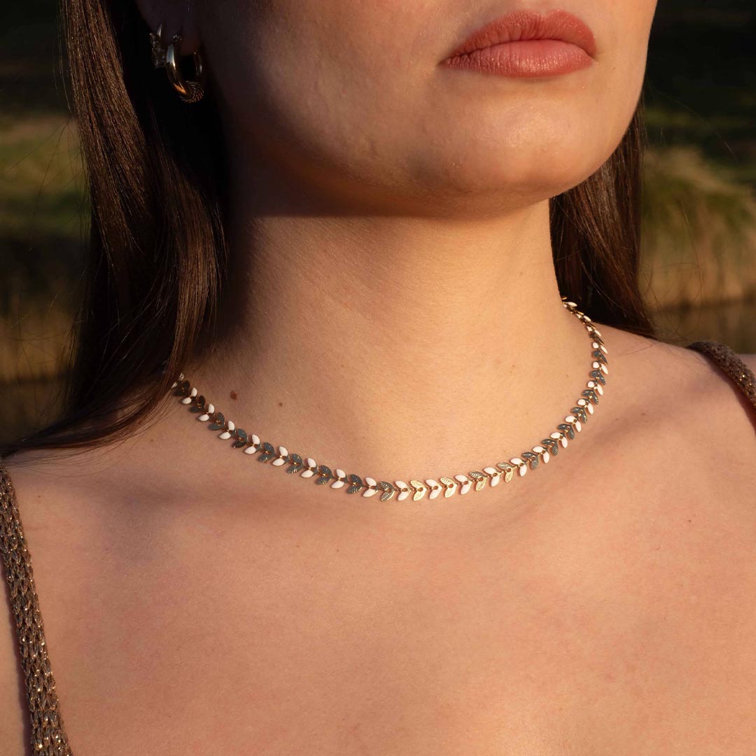 Un magnifique collier en acier inoxydable "Taina" de couleur Or, le bijou possède une chaine en forme de fleur / feuilles alternant les couleurs or et blanc. Le collier est porté par une jolie modèle aux cheveux bruns. On y voit le bas de son visage. La lumière de la photo est chaude.