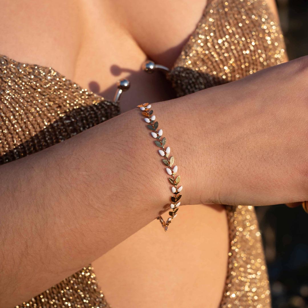 Un magnifique bracelet en acier inoxydable "Taina" de couleur Or, le bijou possède une chaine en forme de fleur / feuilles alternant les couleurs or et blanc. Le bracelet est porté par une modèle avec une robe dorée brillante qui reflète le soleil.