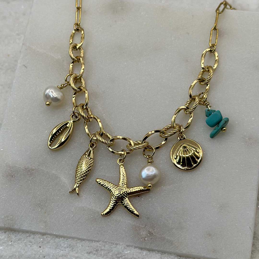 Un magnifique collier en acier inoxydable "Vaia" de couleur Or, le bijou possède plusieurs charms : deux perles blanches, deux coquillage, une étoile de mer et une pierre bleu turquoise, ce bijou est posé sur une plaque de marbre blanche qui elle même est posé sur du sable blanc qui fait office de fond d'image.