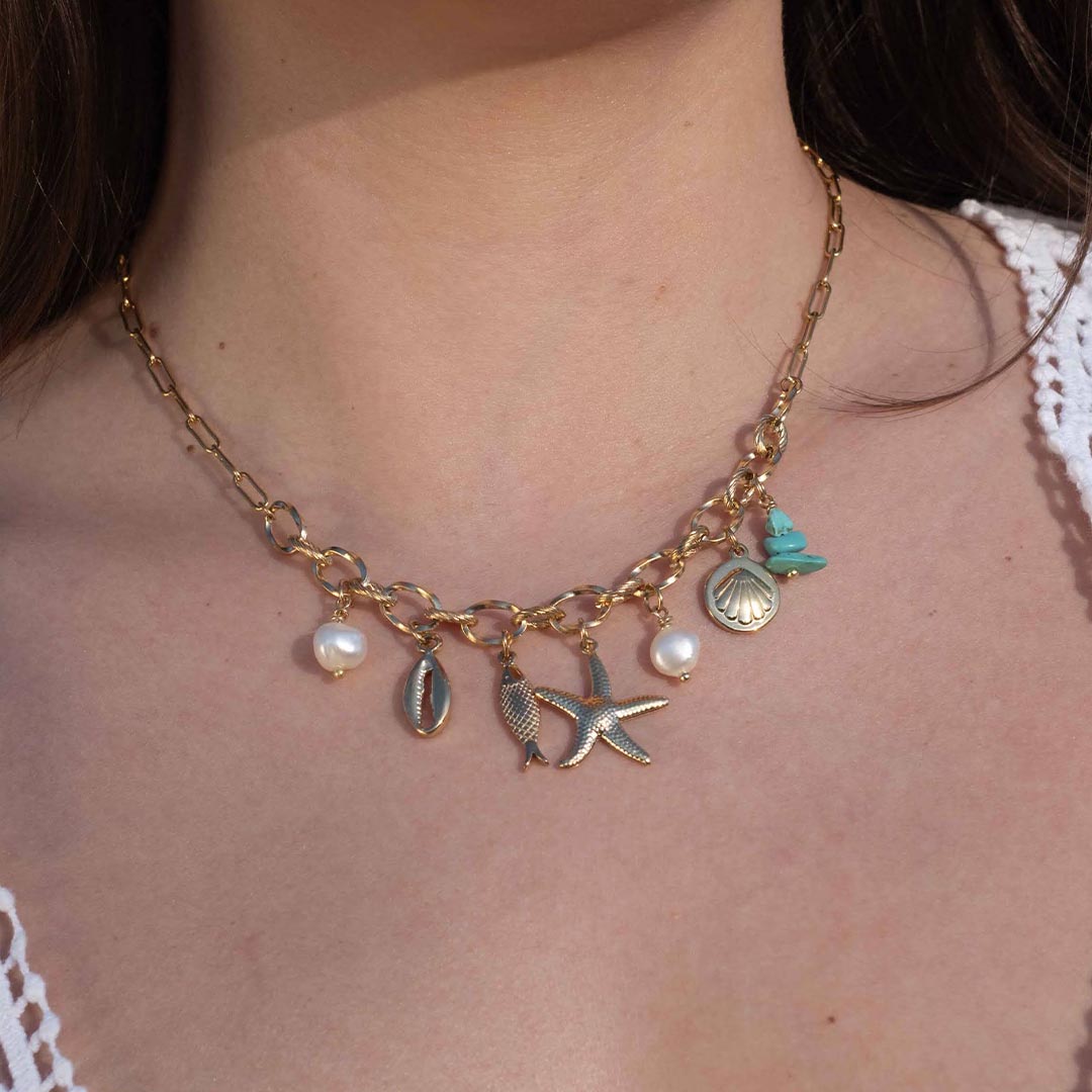 Un magnifique collier en acier inoxydable "Vaia" de couleur Or, le bijou possède plusieurs charms : deux perles blanches, deux coquillage, une étoile de mer et une pierre bleu turquoise. Le collier est porté par une modèle aux cheveux bruns.