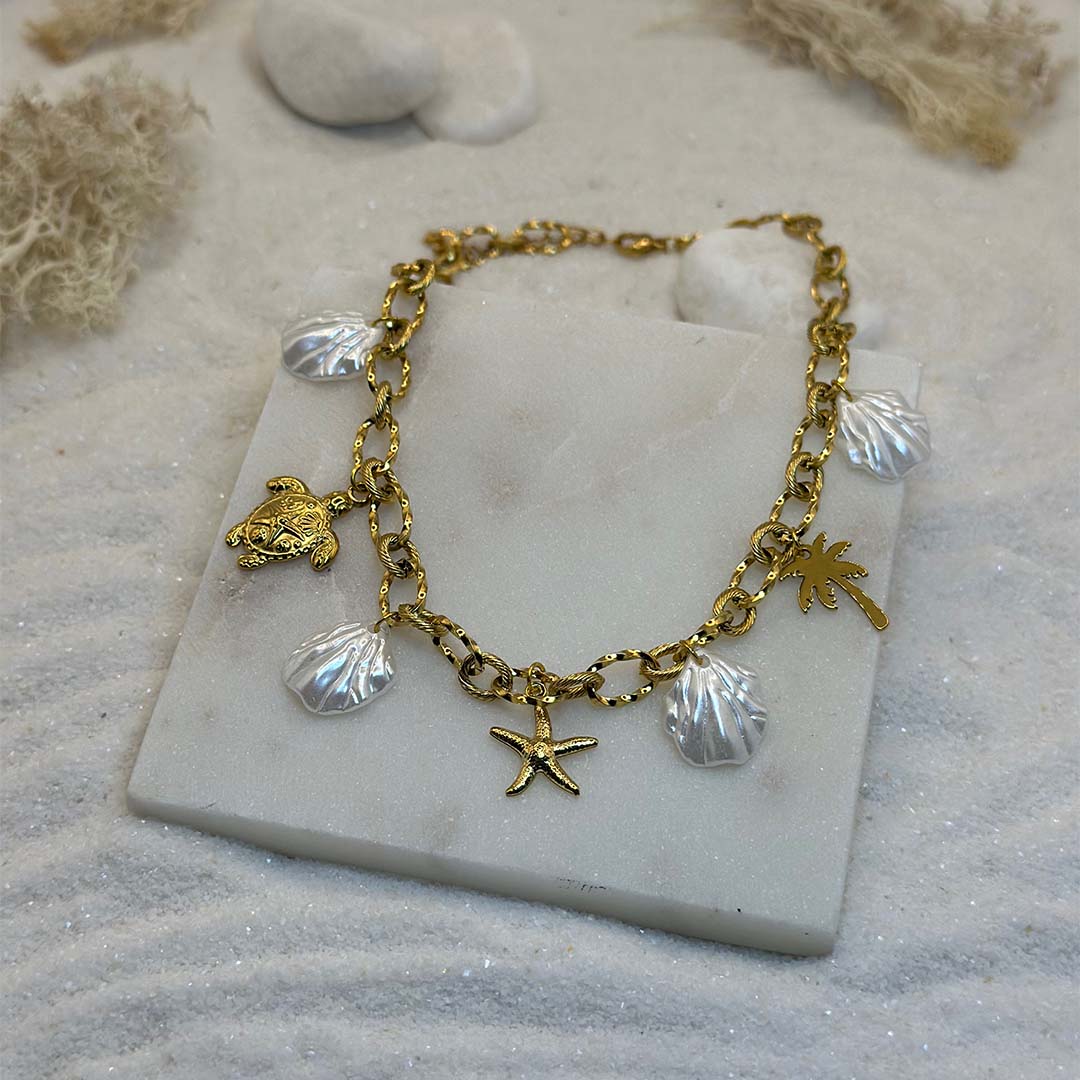 Un magnifique collier en acier inoxydable "Vanea" de couleur Or, le bijou possède plusieurs charms : plusieurs coquillages de couleur blanc, une tortue de couleur or, une étoile de mer de couleur or, un palmier de couleur or, ce bijou est posé sur une plaque de marbre blanche qui elle même est posé sur du sable blanc qui fait office de fond d'image.