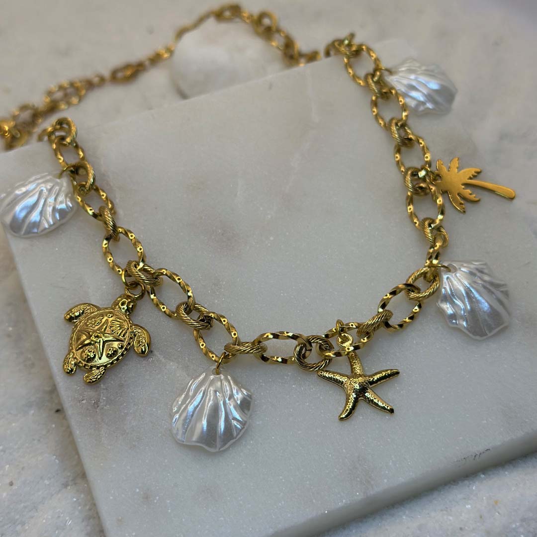 Un magnifique collier en acier inoxydable "Vanea" de couleur Or, le bijou possède plusieurs charms : plusieurs coquillages de couleur blanc, une tortue de couleur or, une étoile de mer de couleur or, un palmier de couleur or, ce bijou est posé sur une plaque de marbre blanche qui elle même est posé sur du sable blanc qui fait office de fond d'image.