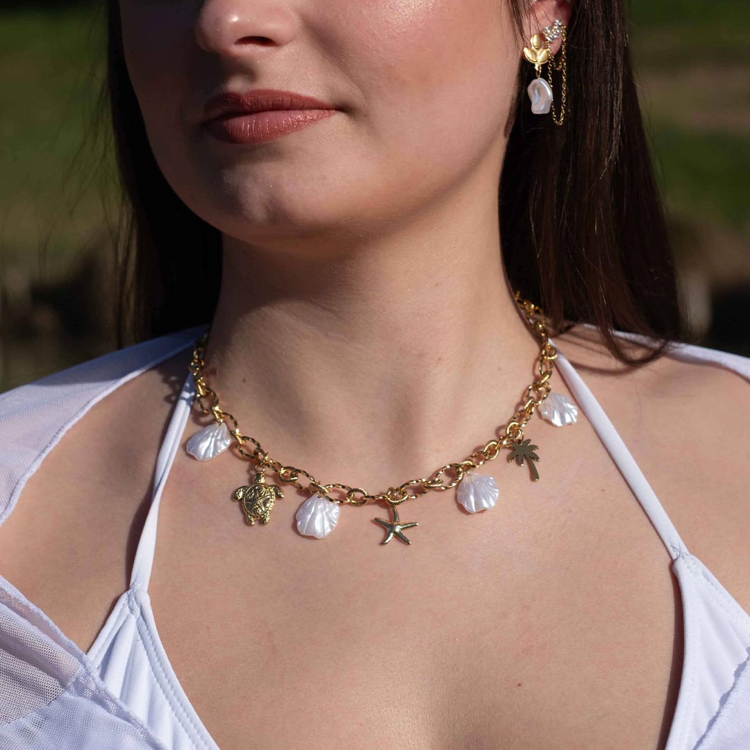 Un magnifique collier en acier inoxydable "Vanea" de couleur Or, le bijou possède plusieurs charms : plusieurs coquillages de couleur blanc, une tortue de couleur or, une étoile de mer de couleur or, un palmier de couleur or. Le collier est porté par une jolie modèle aux cheveux bruns. On y voit le bas de son visage ainsi que son oreille gauche avec les boucles "fidji" et "ezea". La modèle porte des vêtements blancs.