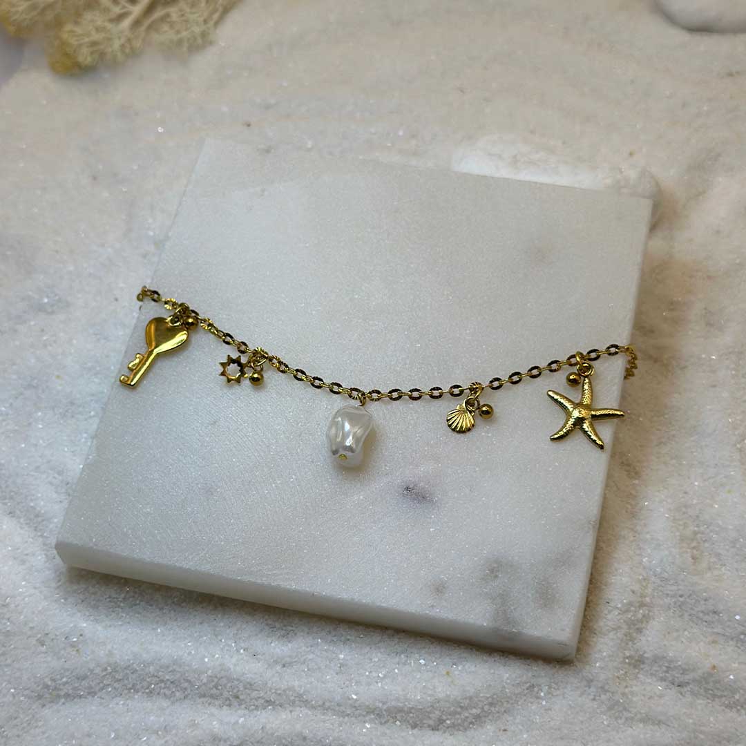 Un magnifique bracelet en acier inoxydable "Vania" de couleur Or, ce bijou possède plusieurs charms : une clef en forme de coeur a la base, un soleil, une étoile de mer, un coquillage ainsi qu'une perle blanche ,le bijou est posé sur une plaque de marbre blanche qui elle même est posé sur du sable blanc qui fait office de fond d'image.