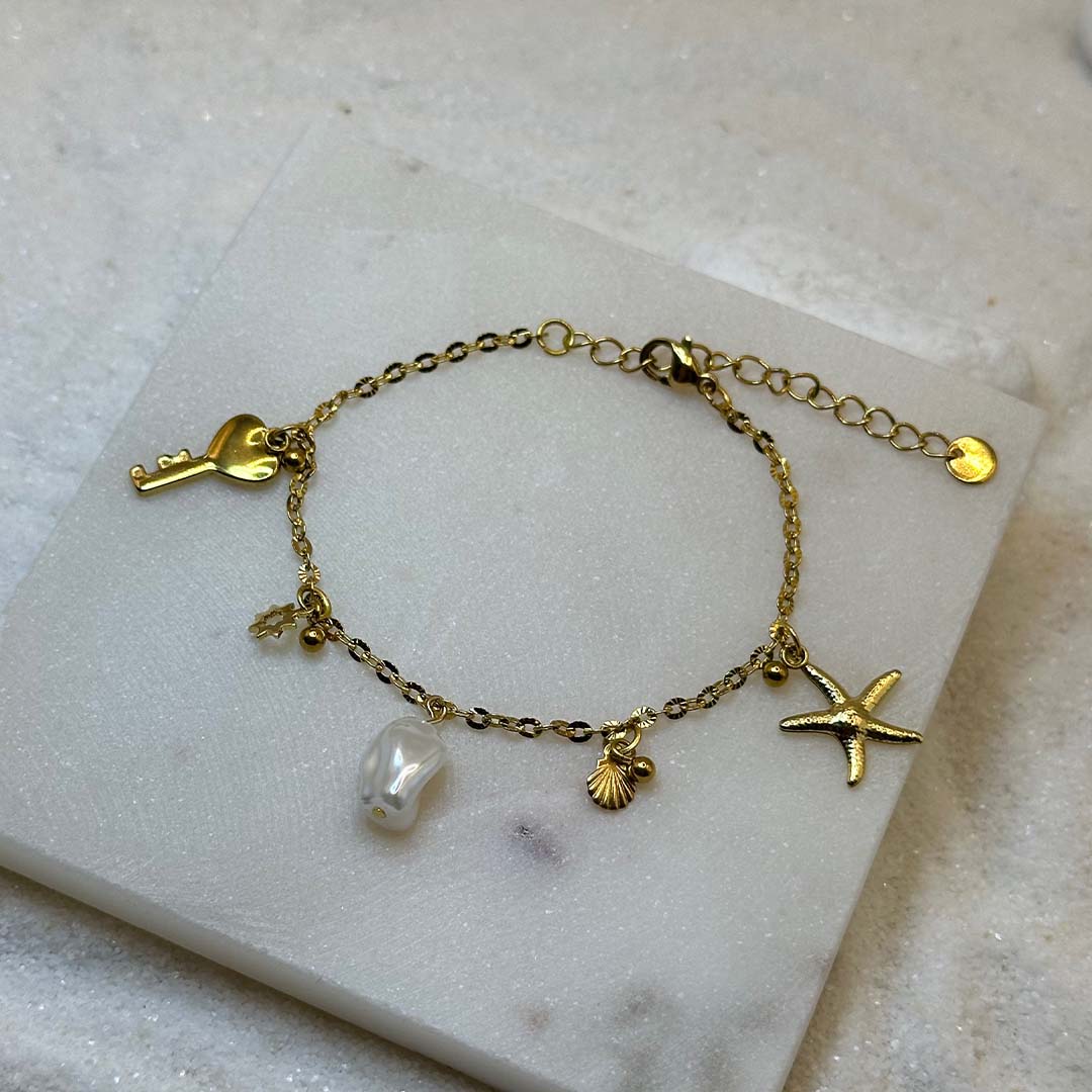 Un magnifique bracelet en acier inoxydable "Vania" de couleur Or, ce bijou possède plusieurs charms : une clef en forme de coeur a la base, un soleil, une étoile de mer, un coquillage ainsi qu'une perle blanche ,le bijou est posé sur une plaque de marbre blanche qui elle même est posé sur du sable blanc qui fait office de fond d'image.