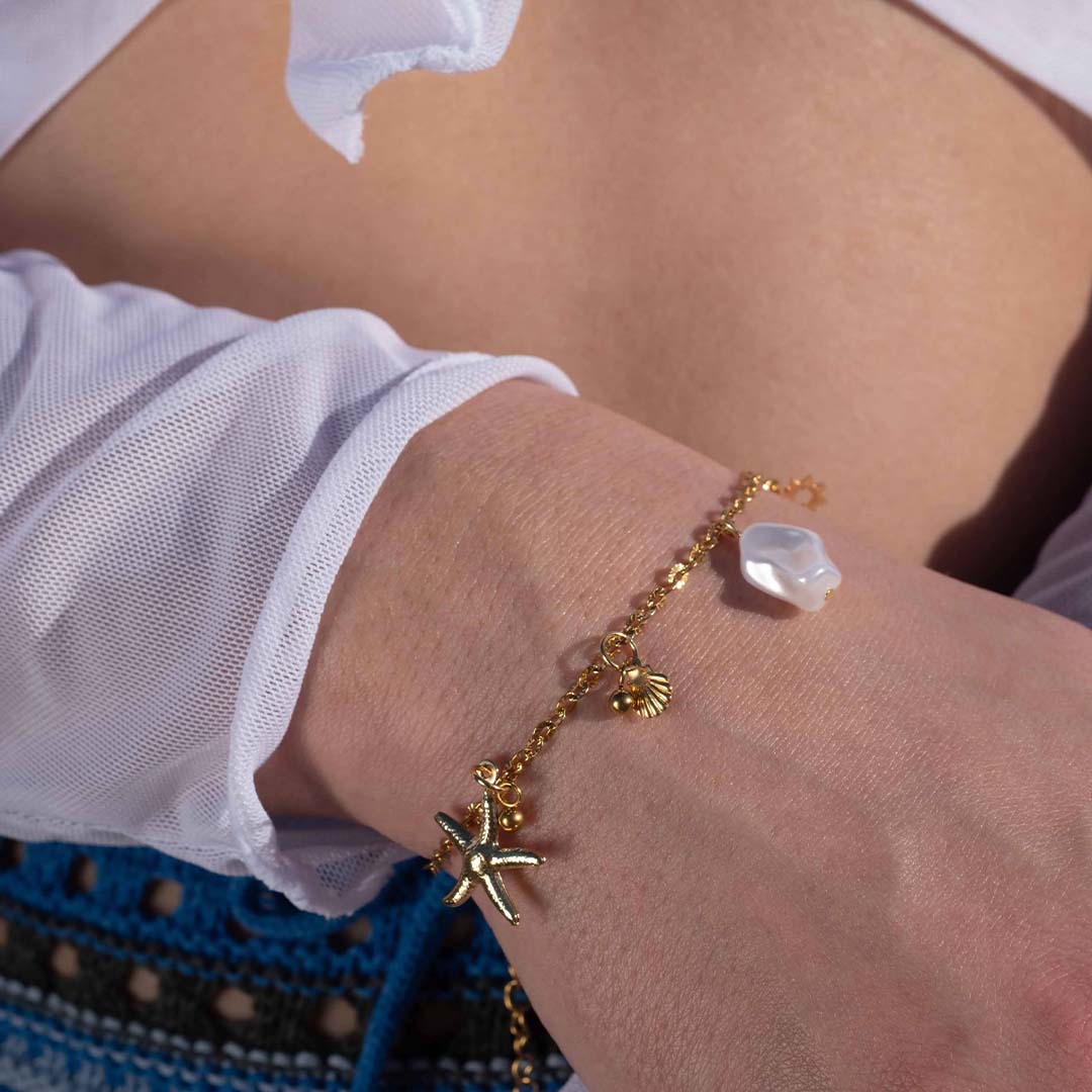 Un magnifique bracelet en acier inoxydable "Vania" de couleur Or, ce bijou possède plusieurs charms : une clef en forme de coeur a la base, un soleil, une étoile de mer, un coquillage ainsi qu'une perle blanche. Le bracelet est porté par une modèle, on y voit son poignet et son ventre en arrière plan.