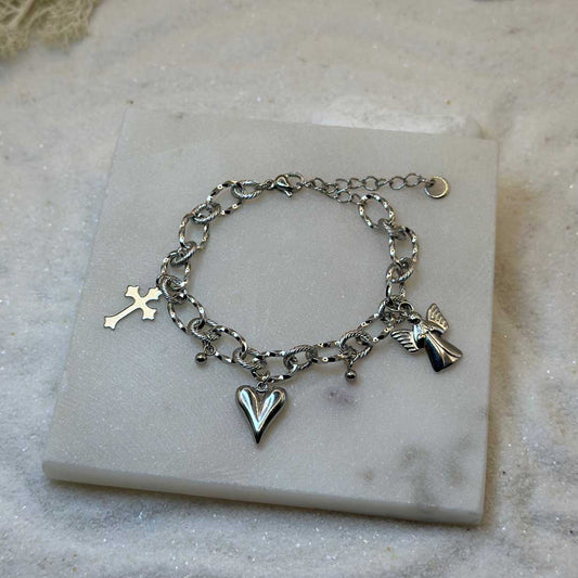 Un magnifique bracelet en acier inoxydable "Angely" est au centre de l'image. Le bijou possède des charmes : un ange, une croix et un coeur. Le bracelet est posé sur une plaque de marbre. Il y a du sable blanc en fond.