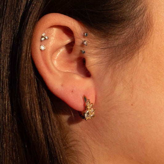 Photo d'une oreille prise de près pour montrer ce magnifique bijou "ania" en acier inoxydable qui est une boucle d'oreille avec des feuilles en strass. Il est de couleur doré / or. Sur l'oreille on retrouve aussi des piercing. La lumière de la photo est chaude.