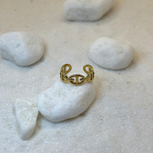 Une très jolie bague en or "ari" est au centre de l'image, posée sur un caillou blanc. La bague est sur le dessus de la pierre. Le bijoux en acier inoxydable est en forme de maillons. Tout autour se trouve du sable blanc et d'autres cailloux.