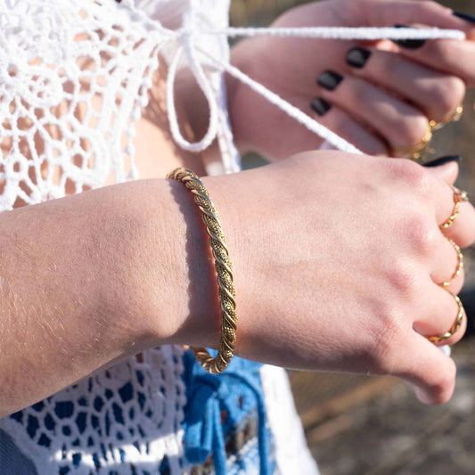Photo d'un poignet pris de près pour montrer ce magnifique bijou "atilia" en acier inoxydable qui est un bracelet / un jonc torsadé / tressé. Il est de couleur doré / or. En arrière plan nous pouvons voir les mains de la modèle.