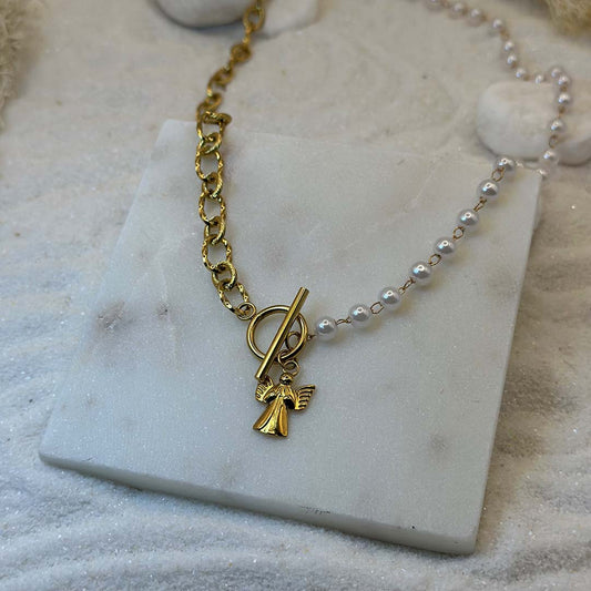 Un très beau collier de deux matières se trouve au centre de l'image. La partie gauche est en or, et la partie droite en perles. Au centre se trouve un charms en or : un ange. Le bijoux en acier inoxydable est posé sur une plaque de marbre. Autour se trouvent des cailloux et du sable blanc.