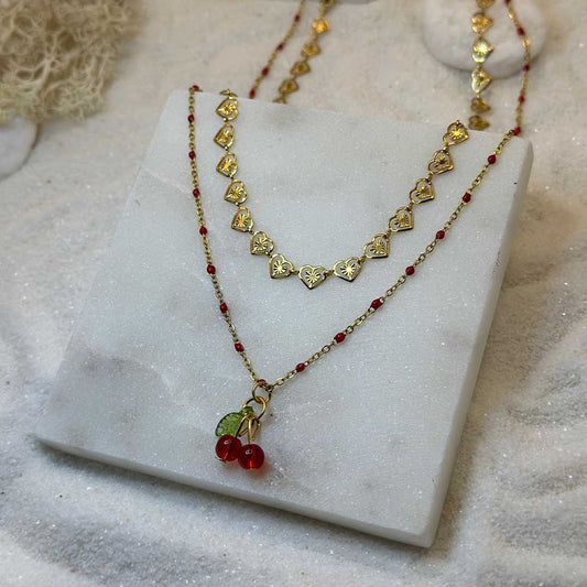 Un magnifique collier en or "cerise" est au centre de l'image, joliment posé sur une plaque de marbre vu d'en haut. Le bijoux en acier inoxydable est séparé en deux parties : l'une est faite de coeur en or. L'autre d'une chaîne en or, avec des jointures rouge, ainsi que d'un charms en cerises. Autour se trouve du sable blanc.