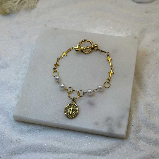Un magnifique collier en or "charlie" est au centre de l'image. C'est un très beau bracelet en acier inoxydable composé de perles, de croix, de maillons, ainsi que d'un pendentif ovale avec une croix. Le bijoux est posé sur une plaque de marbre. Tout autour se trouve du sable blanc.