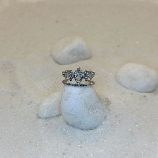 Une très jolie bague en argent "doli" est au centre de l'image, posée sur un petit cailloux blanc. Le bijoux est en acier inoxydable, et ressemble à une couronne. Autour se trouvent d'autres cailloux et du sable blanc.