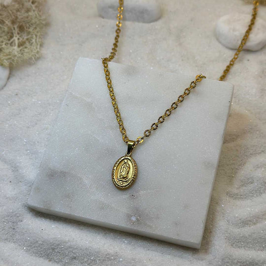Un très jolie collier en or "eden" est au centre de l'image, posé sur une plaque de marbre. Le bijoux en acier inoxydable ressemble à un collier chrétien, avec un pendentif et d'un saint. Autour se trouve du sable blanc.