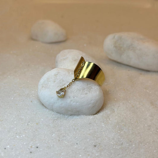 Une très jollie bague en or "enea" est au centre de l'image, posée sur un cailloux blanc en gros plan. Le bijoux en acier inoxydable est en deux parties : la première est une base en or, en forme de flèche. La seconde est un strass, relié par une chaîne en or à la base. Autour se trouve du sable blanc.