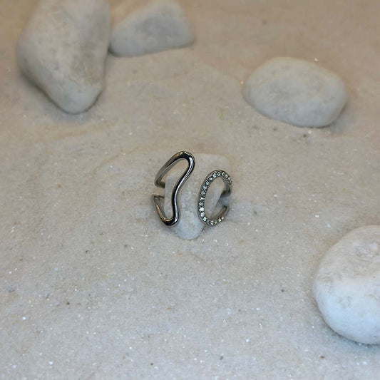 Une magnifique bague en argent "era" est au centre de l'image, posées sur du sable et blanc et entourant un caillou blanc. Le bijoux en acier inoxydable possède des strass sur sa partie droite. Autour se trouvent d'autres cailloux et du sable.