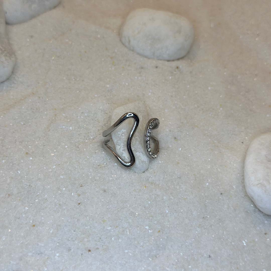 Une magnifique bague en argent "era" est au centre de l'image, posées sur du sable et blanc et entourant un caillou blanc. Le bijoux en acier inoxydable possède des strass sur sa partie droite, et nous le voyons de 3/4. Autour se trouvent d'autres cailloux et du sable.