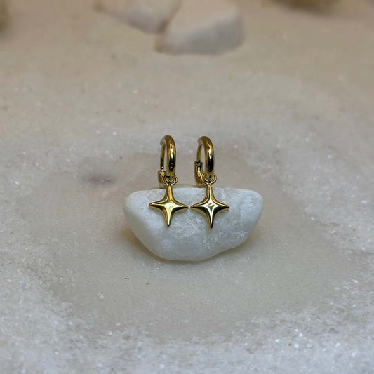 Deux très belles boucles d'oreilles en or "estella", sont au centre de l'image, posées sur un cailloux blanc et vu de face. Ce sont des bijoux en acier inoxydable en forme d'étoiles. Tout autour se trouve du sable blanc.