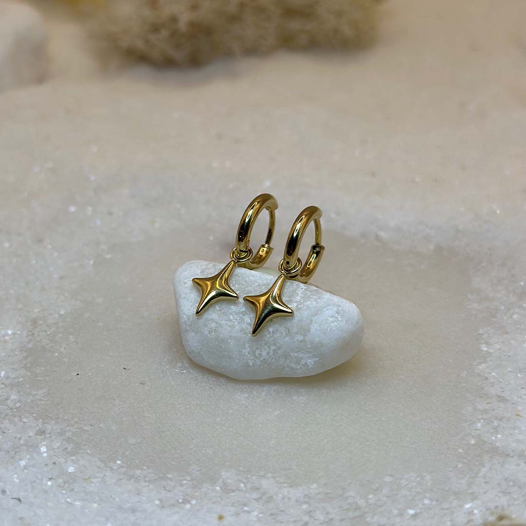 Deux très belles boucles d'oreilles en or "estella", sont au centre de l'image, posées sur un cailloux blanc et vu de 3/4. Ce sont des bijoux en acier inoxydable en forme d'étoiles. Tout autour se trouve du sable blanc.