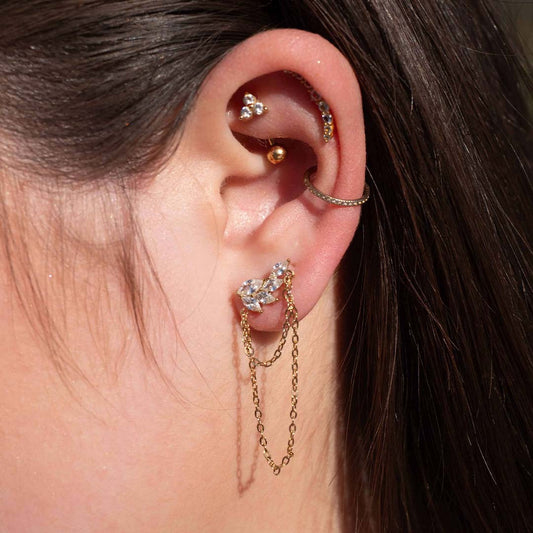 Photo d'une oreille prise de près pour montrer ce magnifique bijou "ezea" en acier inoxydable qui est une boucle d'oreille dorée avec des feuilles en strass et une double chaînette. En arrière plan nous pouvons voir les cheveux bruns de la modèle ainsi que des piercings.