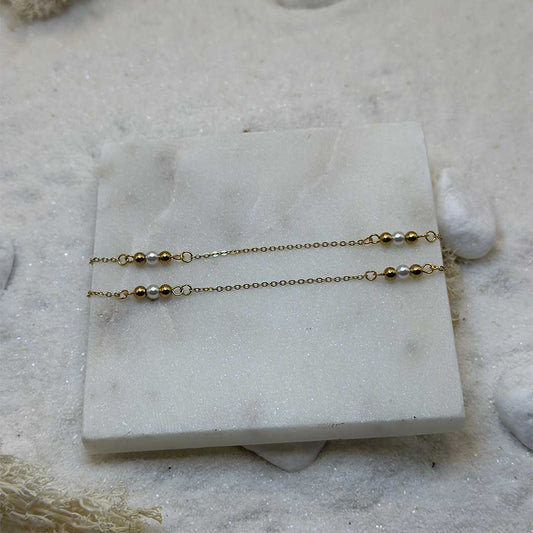 Une très belle chaîne de taille en or "felicie" est au centre de l'image. Le bijoux en acier inoxydable possède des perles en or et perles basiques, les maillons sont en or. Les deux chaînes sont posées sur une plaque de marbre. Tout autour se trouve du sable blanc, des cailloux et des algues.