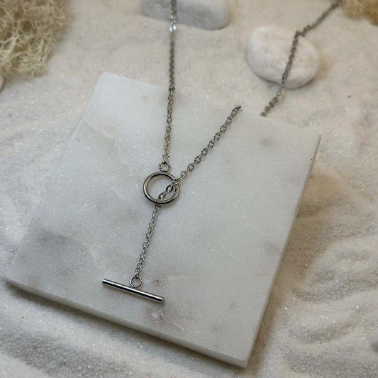 Un magnifique collier en argent "fenicie" est au centre de l'image, posé sur une plaque en marbre. Le bijoux en acier inoxydable est composé d'une chaîne en forme de maillons étoilés, ainsi que d'un cercle et d'une barre lisses. En fond se trouve du sable blanc.