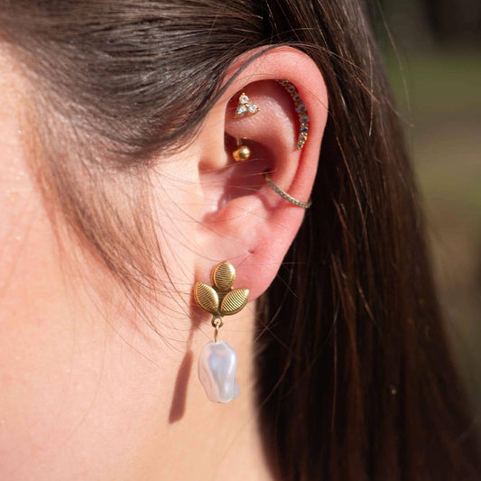 Photo d'une oreille prise de près pour montrer ce magnifique bijou "fidji" en acier inoxydable qui est une boucle d'oreille avec des feuilles dorées texturées auquel est accroché une perle nacrée blanche. En arrière plan nous pouvons voir les cheveux bruns de la modèle ainsi de des piercings.