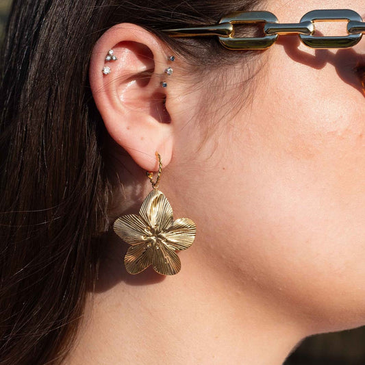 Photo d'une oreille prise de près pour montrer ce magnifique bijou "flora" en acier inoxydable qui est une boucle d'oreille avec un anneau torsadé portant une fleur style hibiscus texturée de couleur or / doré. En arrière plan nous pouvons voir les cheveux bruns de la modèle ainsi de des piercings et une branche de lunette.