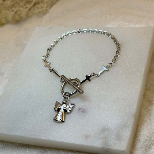 Un magnifique bracelet en acier inoxydable "Guillia" en couleur argent, ce bijoux possède deux différentes chaine, une en forme de croix et l'autre avec des maillons fins, il possède aussi un pendentifs en forme d'ange de couleur argent, le bijoux est posé sur une plaque de marbre blanche et est elle même posé sur du sable blanc qui fait office de fond d'image.