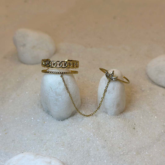 Une magnifique bague en or, en deux parties, "inicia" est au centre de l'image, posée sur deux cailloux blancs. Le bijoux en acier inoxydable est composé d'une première bague en forme de maillons, reliée par une chaîne à une seconde bague avec un design en forme de fleur. Autour se trouve du sable blanc.
