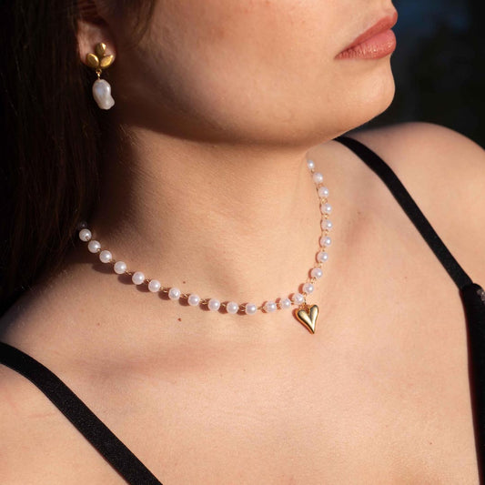 Ce magnifique bijou "Jane" en acier inoxydable est un collier de parles blanches avec un pendentif coeur de couleur or. Il est porté par une modèle dont on voit le bas du visage aisni que son oreille et ses cheveux bruns.
