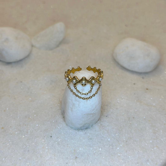 Une très belle bague en or "livia" est posée autour d'un caillou blanc, au centre de l'image. Le bijoux en acier inoxydable est aussi composé de 2 chaînes en or, reliées à des perles. Tout autour se trouve du sable blanc.