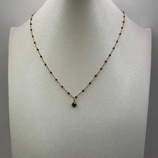 Ce joli bijou "Lola" en acier inoxydable est un collier avec une chaîne dorée et boules noires. Il est composé d'un petit pendentif noir brillant à strass. Il est posé sur un buste blanc pailleté qui est lui même posé devant un fond blanc.
