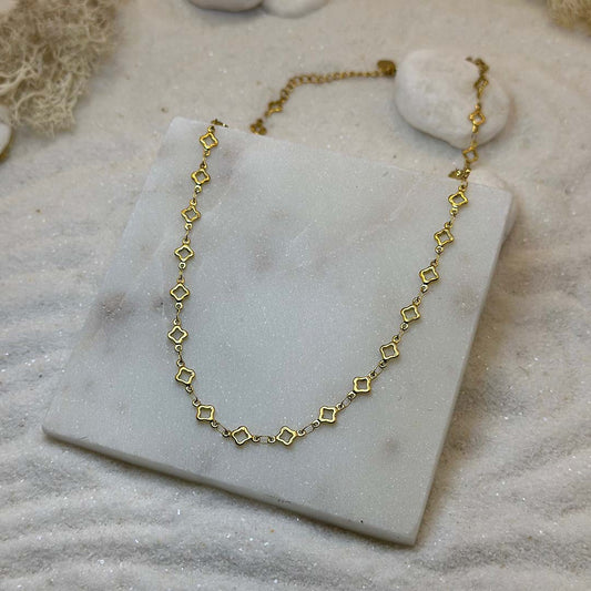 Un magnifique collier en or "lori" est au centre de l'image, posé sur une plaque de marbre. Le bijoux en acier inoxydable est composé d'une chaîne en or, ainsi que des motifs en forme d'étoiles. Autour se trouve du sable blanc.