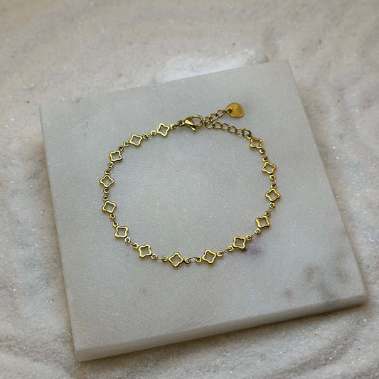 Un magnifique bracelet en or "lori" est au centre de l'image, posé sur une plaque de marbre. Le bijoux en acier inoxydable est composé d'une chaîne en or, ainsi que des motifs en forme d'étoiles. Autour se trouve du sable blanc.