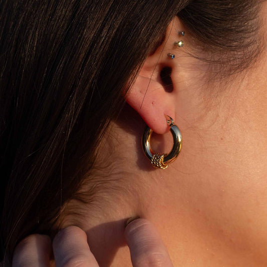 Une modèle porte une magnifique paire de boucles d'oreille en or "lou". Les bijoux en acier inoxydable sont vu de face, composés d'une base lisse et de 3 cordes en relief au centre. La lumière de la photo est chaude. On retrouve en arrière plan les cheveux bruns de la modèle ainsi que des piercings.