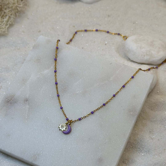 Un magnifique collier en acier inoxydable "Lunicia" de couleur or et violet, ce bijou possède pendentifs lune et soleils de couleurs or et violet ainsi que plusieurs perles violettes tout le long de la chaine, ce bijou est posé sur une plaque de marbre blanche qui elle même est posé sur du sable blanc qui fait office de fond d'image.
