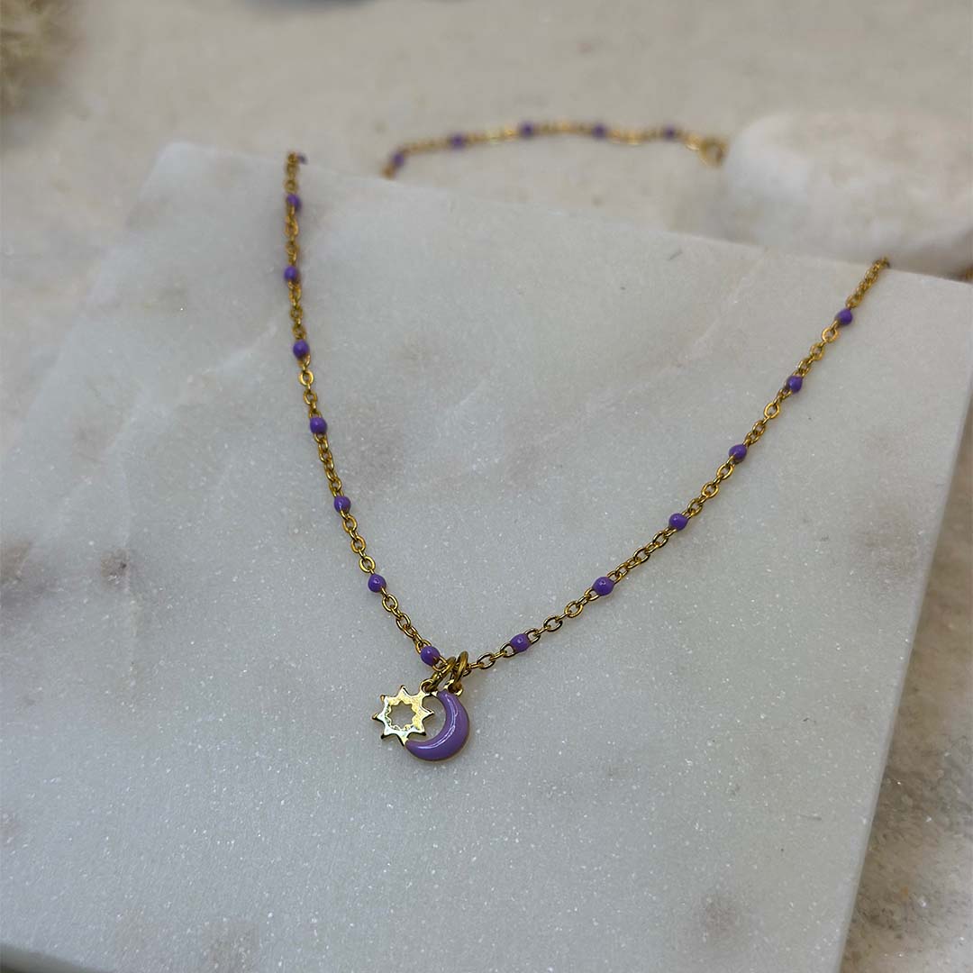 Un magnifique collier en acier inoxydable "Lunicia" de couleur or et violet, ce bijou possède pendentifs lune et soleils de couleurs or et violet ainsi que plusieurs perles violettes tout le long de la chaine, ce bijou est posé sur une plaque de marbre blanche qui elle même est posé sur du sable blanc qui fait office de fond d'image.