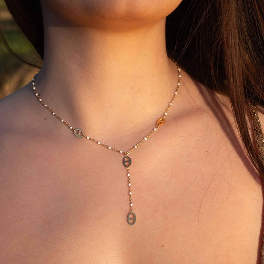 Un magnifique collier en acier inoxydable "Mada" de couleur or et et blanc, ce bijou possède plusieurs pendentifs de forme noeuds marin et des perles blanches  tout le long de la chaine, ce bijou est porté par une modèle brune. La lumière de la photo est chaude.
