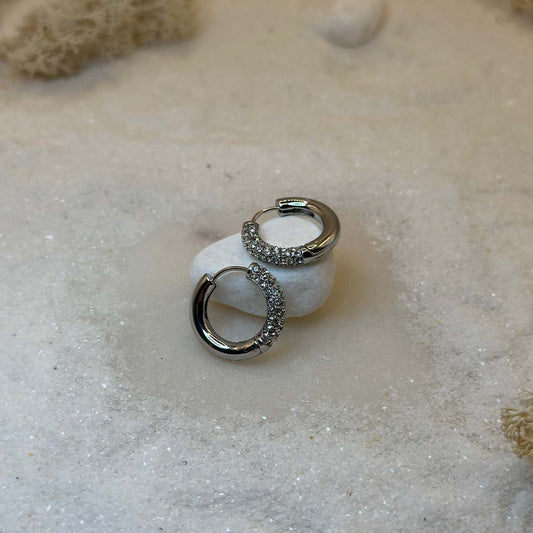 Une magnifique paire de boucle d'oreilles en acier inoxydable "Mandy" de couleur argent, ces boucles d'oreilles sont de forme ronde avec un coté en strass tte bagues scindé en deux, ce bijou est posé sur un caillou blanc qui est posé sur du sable blanc qui fait office de fond d'image.