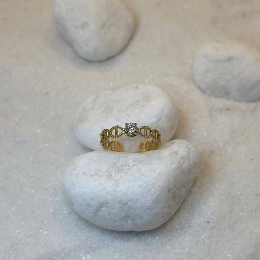 Une magnifique bague en acier inoxydable "Mira" de couleur or, cette bague est de forme ronde avec des noeud marin sur tout le tour et avec un strass au centre, le bijou est posé sur un caillou blanc qui est posé sur du sable blanc qui fait office de fond d'image.